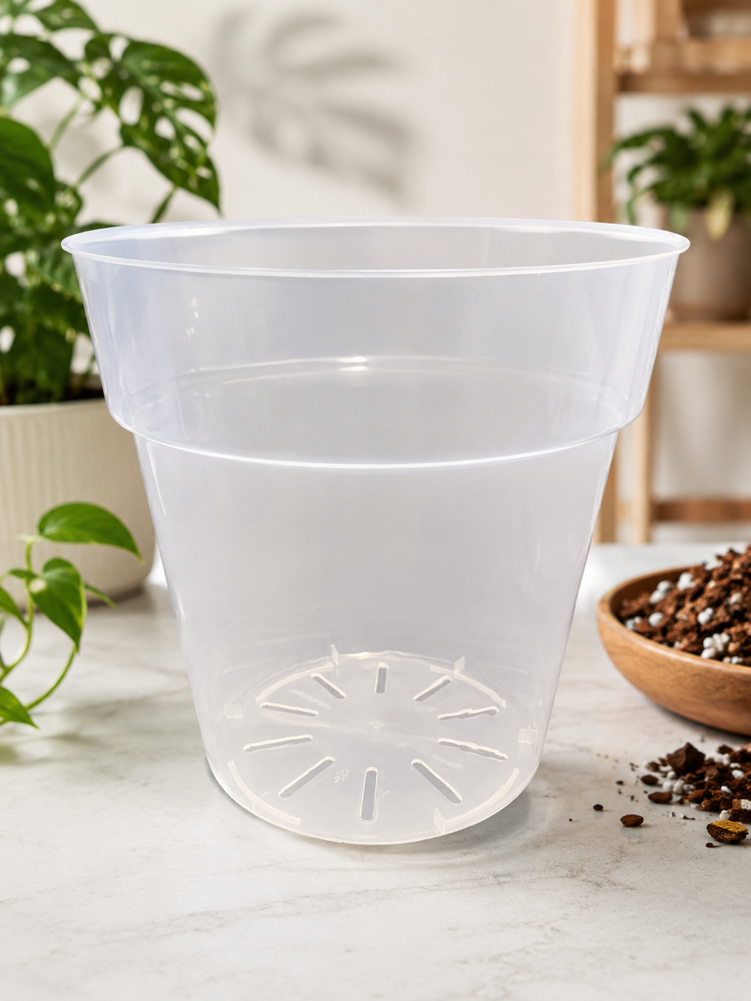 A clear plastic two-tier planter pot with a drainage hole at the bottom, displayed on a light marble surface with houseplants and soil in the background.