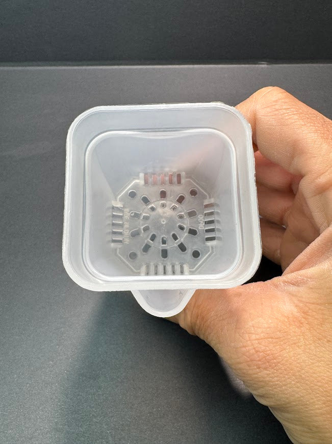 A hand holding a small translucent white plastic square container with a circular filter or strainer insert inside, shown against a dark gray background.