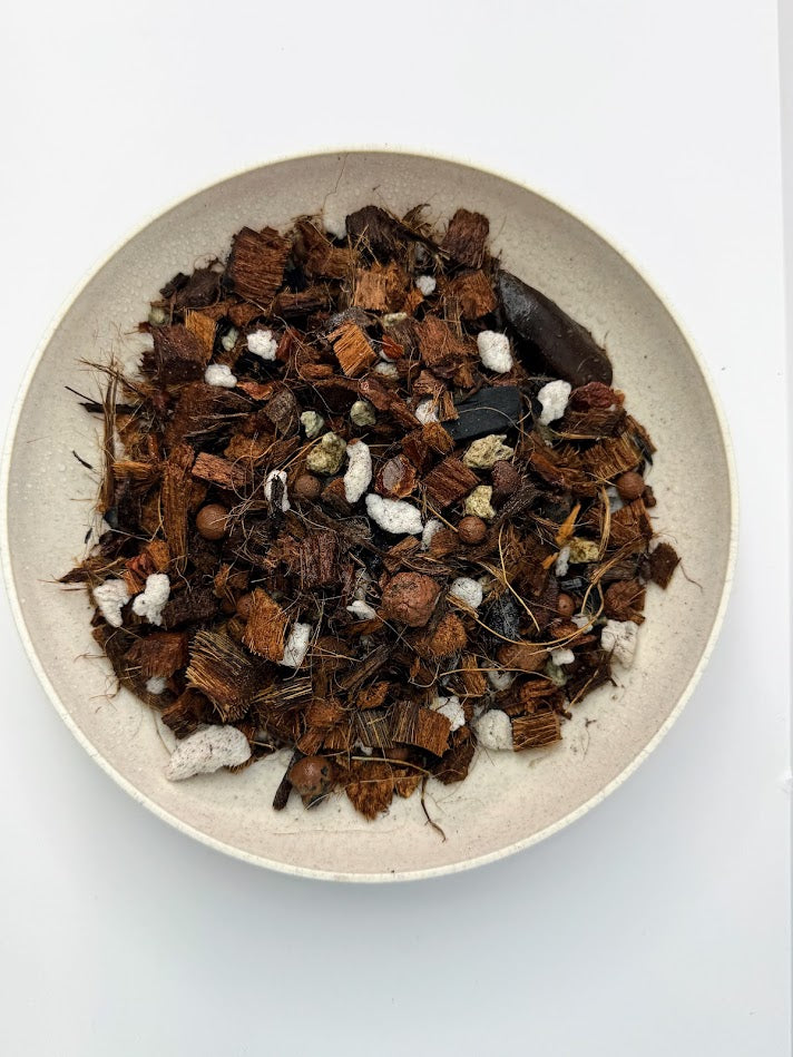 A top-down view of Happy Houseplant Haven Super Chunky Orchid Mix containing brown bark pieces, white perlite, and dark charcoal chunks, presented in a light beige ceramic bowl on a white background.